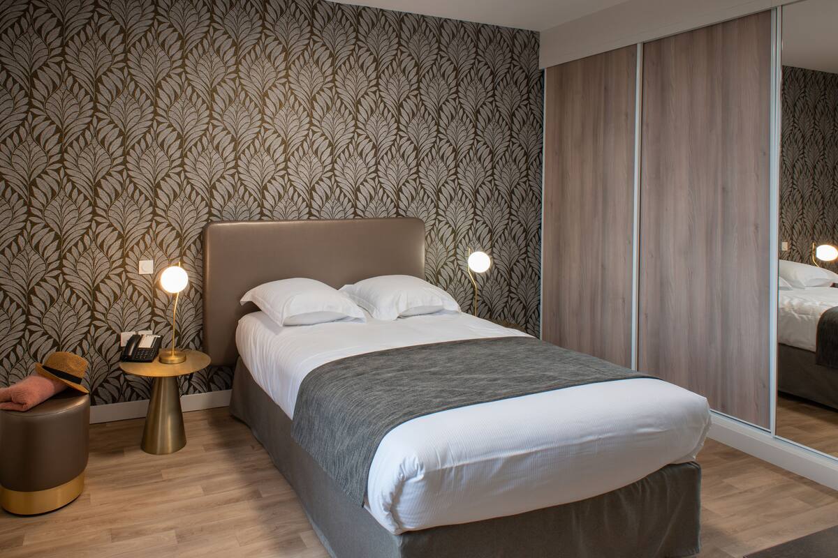 A neatly made bed with a grey blanket, a bedside lamp, and a telephone on a small round table beside it.