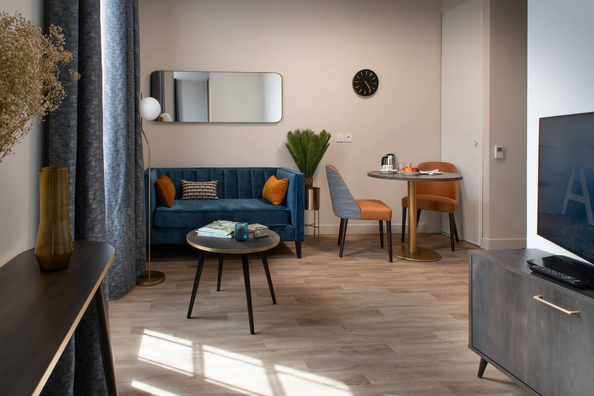 A modern living room with a blue sofa, a round coffee table, a TV, and a dining area with a small table and chairs.