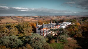 A large, historic building with multiple towers, surrounded by dense trees and a hilly landscape in the background.