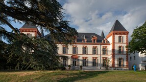 A large, historic building with multiple towers and arched windows, surrounded by a well-maintained lawn and trees.