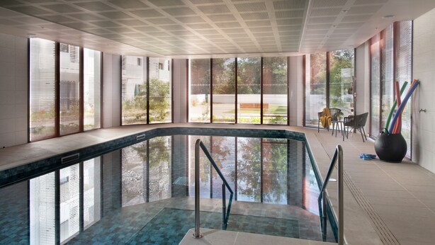 An indoor swimming pool with clear glass walls, a tiled floor, and a ceiling with a grid pattern.