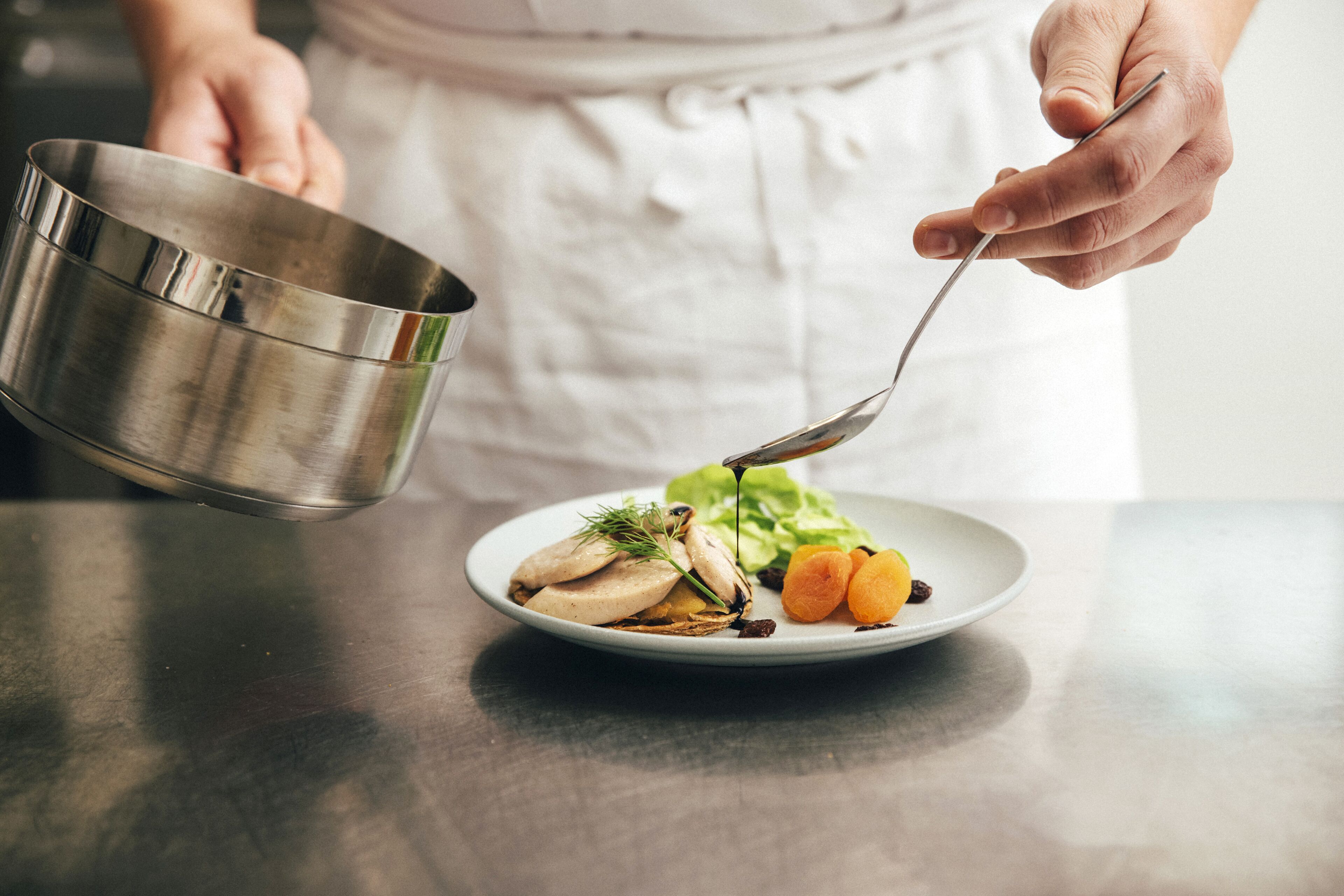 Una persona con delantal blanco sostiene un recipiente metálico y una cuchara sobre un plato con rodajas de champiñones, lechuga y albaricoques.