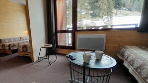 Una habitación acogedora con detalles de madera, una pequeña mesa con dos tazas, una silla, un sofá y una ventana con vistas a los árboles.