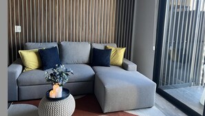 Ein modernes Wohnzimmer mit einem grauen Ecksofa, einem runden Beistelltisch mit Kerzen und einer Wand mit Holzlamellen.