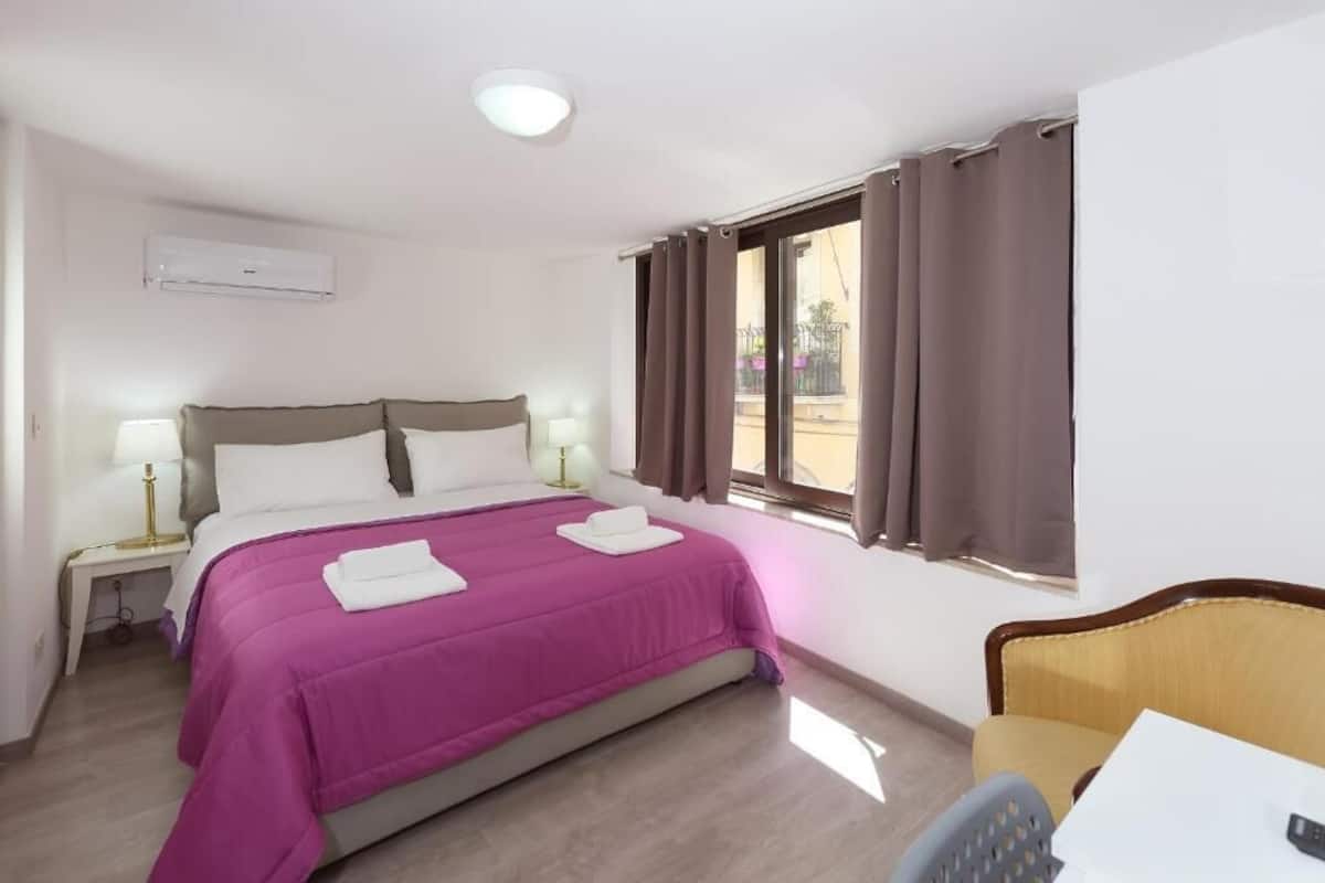 A hotel room with a bed, bedside lamps, a window with curtains, a chair, and a desk with a lamp.