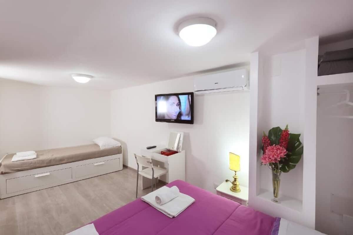 A compact hotel room with a bed, a desk, a chair, a TV, a lamp, and a vase with flowers.