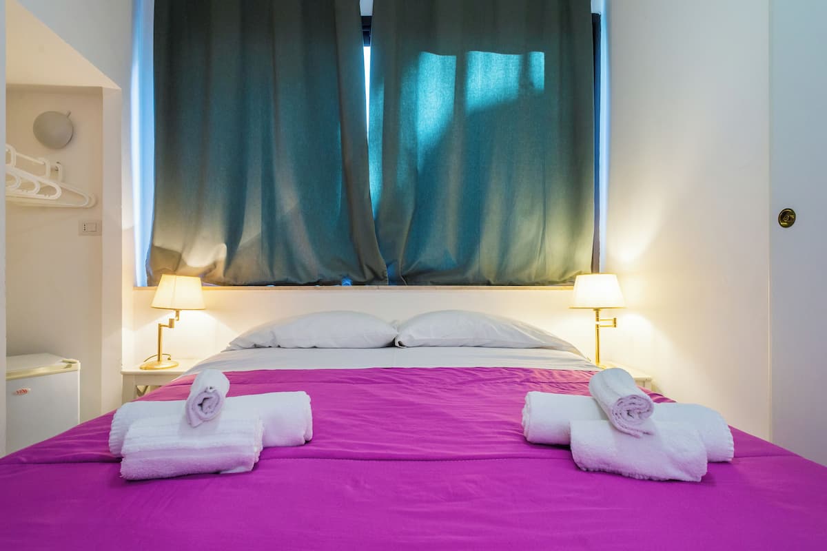 A hotel room with a bed, two bedside lamps, a window with curtains, and two towels on the bed.