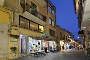 A street view at dusk with a brightly lit storefront, outdoor seating, and a building with balconies. - Casa Fresta (Taormina)