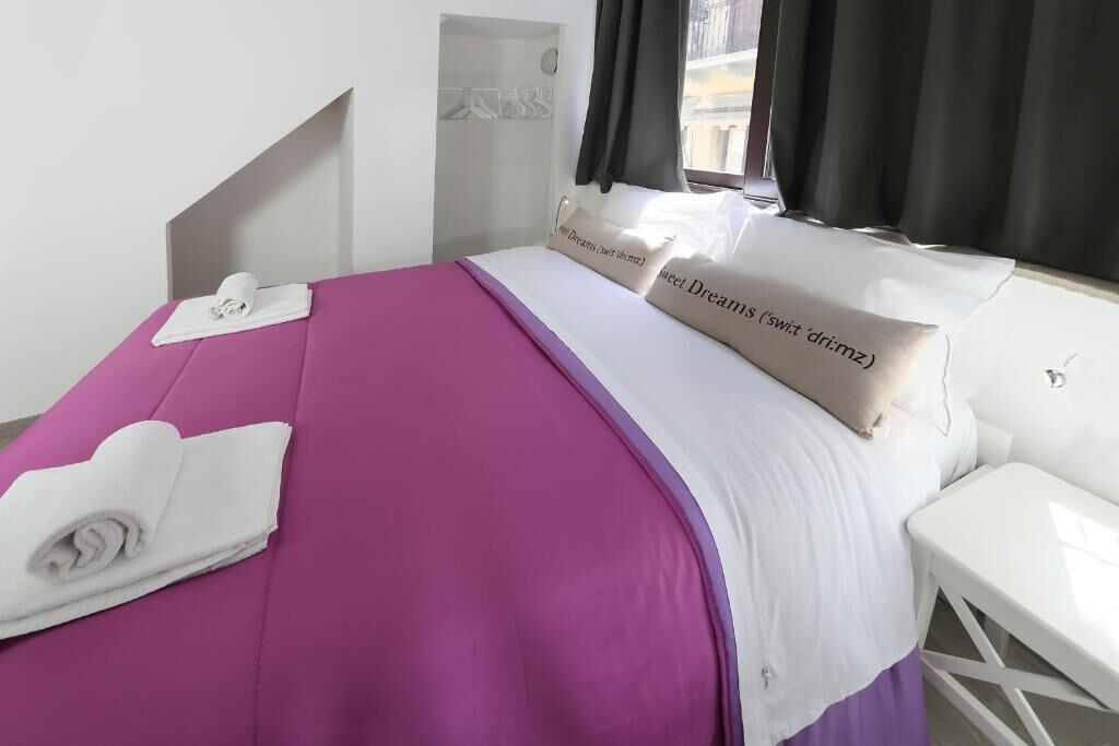 Un lit bien fait, avec une couvre-lit violette et du linge de lit blanc, une table de chevet blanche et une fenêtre ornée de rideaux noirs.