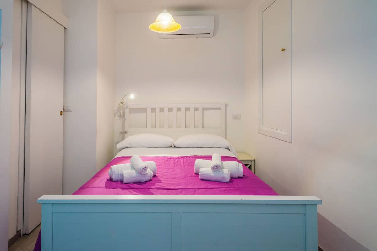 A bedroom with a bed, white headboard, two pillows, a nightstand, and a wall-mounted air conditioner.