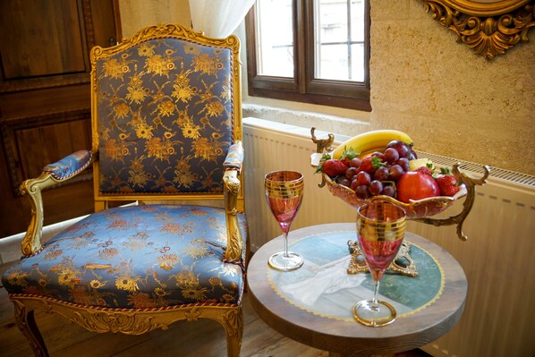 A room with a blue upholstered chair with gold trim, a round table with a fruit bowl and two glasses, and a window with a view of the outside. - Yesil Konak Cave Hotel (Ürgüp)