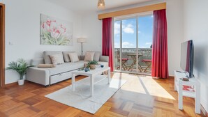 A modern living room with a sofa, coffee table, and a TV. There is a balcony with red curtains and a view of the sky. - Camara de Lobos Apartment (Camara de Lobos)