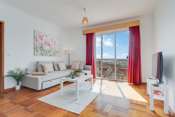 Una sala moderna con sofá, mesa de centro y televisor. Hay un balcón con cortinas rojas y vista al cielo.