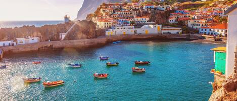 Un pueblo costero con edificios en un acantilado con vista al mar, barcos en el agua y un faro a lo lejos.