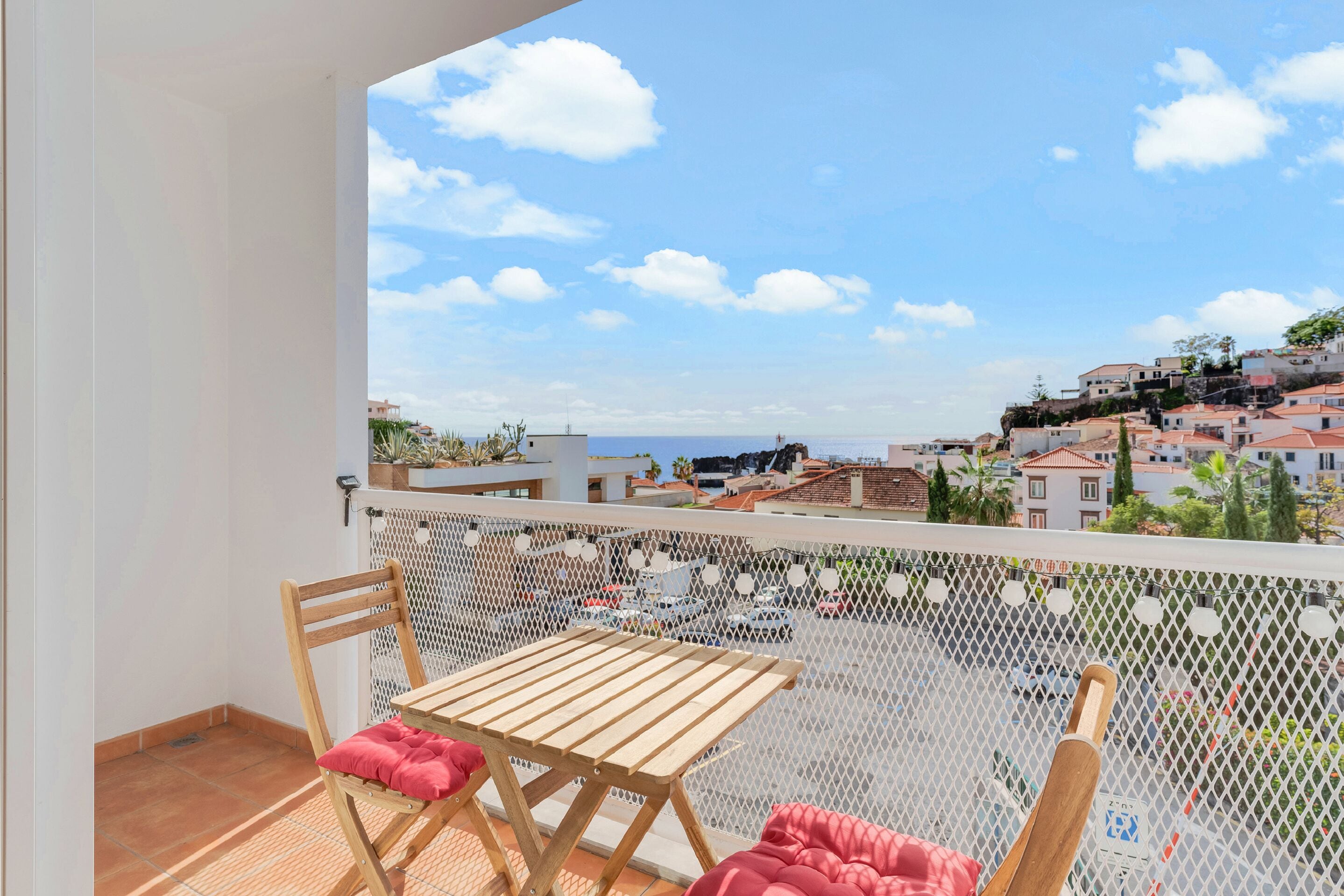 Un balcon avec une table et des chaises en bois, une barrière en treillis blanc et une vue sur une ville côtière.