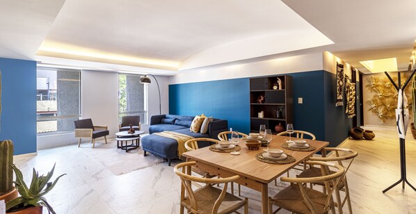 Ein modernes Wohnzimmer mit blauer Akzentwand, einer Couch, einem Esstisch für vier Personen und einem Bücherregal.