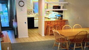 Un coin repas avec une table et des chaises en bois, une cuisine à l’arrière-plan et une horloge au mur.