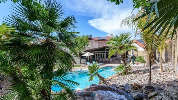A pool area with palm trees, a stone waterfall feature, and a building with a red-tiled roof in the background. - Nomad Gamer Pad- A Mountain Reserve Home w/ Pool, Spa, Esports Arena & Squat Gym (Phoenix)
