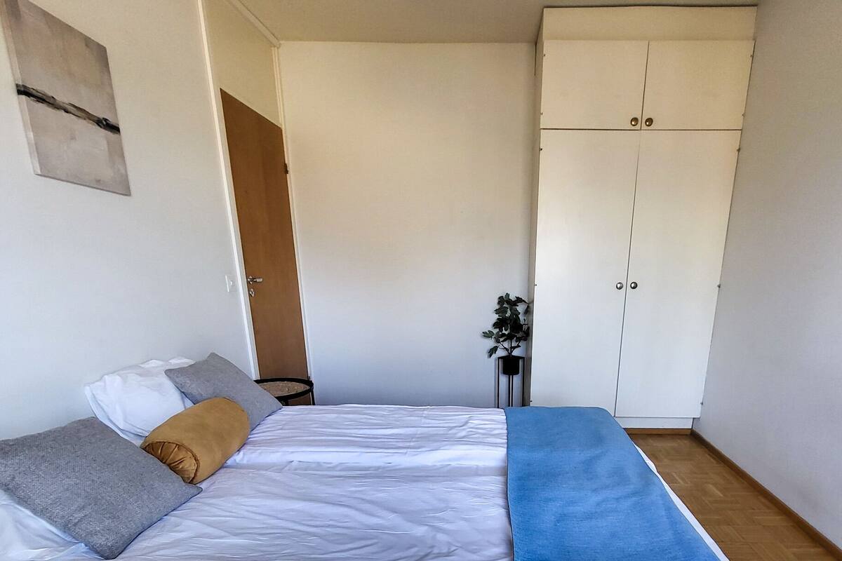 Ein Schlafzimmer mit einem Bett, einer blauen Decke, Holzboden, einem Kleiderschrank und einer Pflanze.