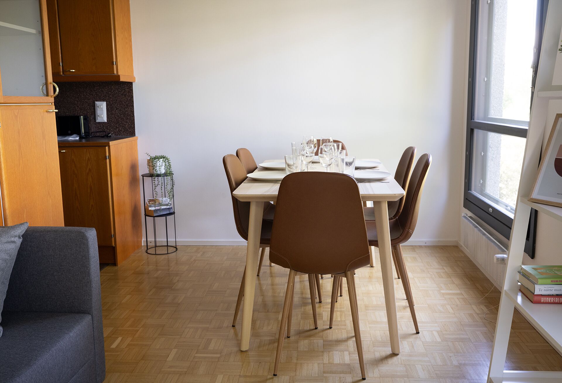 A dining area with a wooden table set for four, surrounded by brown chairs. A small plant is on a metal stand beside the table.