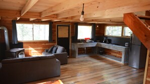 Interior de cabana com chão de madeira, sofá, mesa de jantar, cozinha com geladeira e escada.