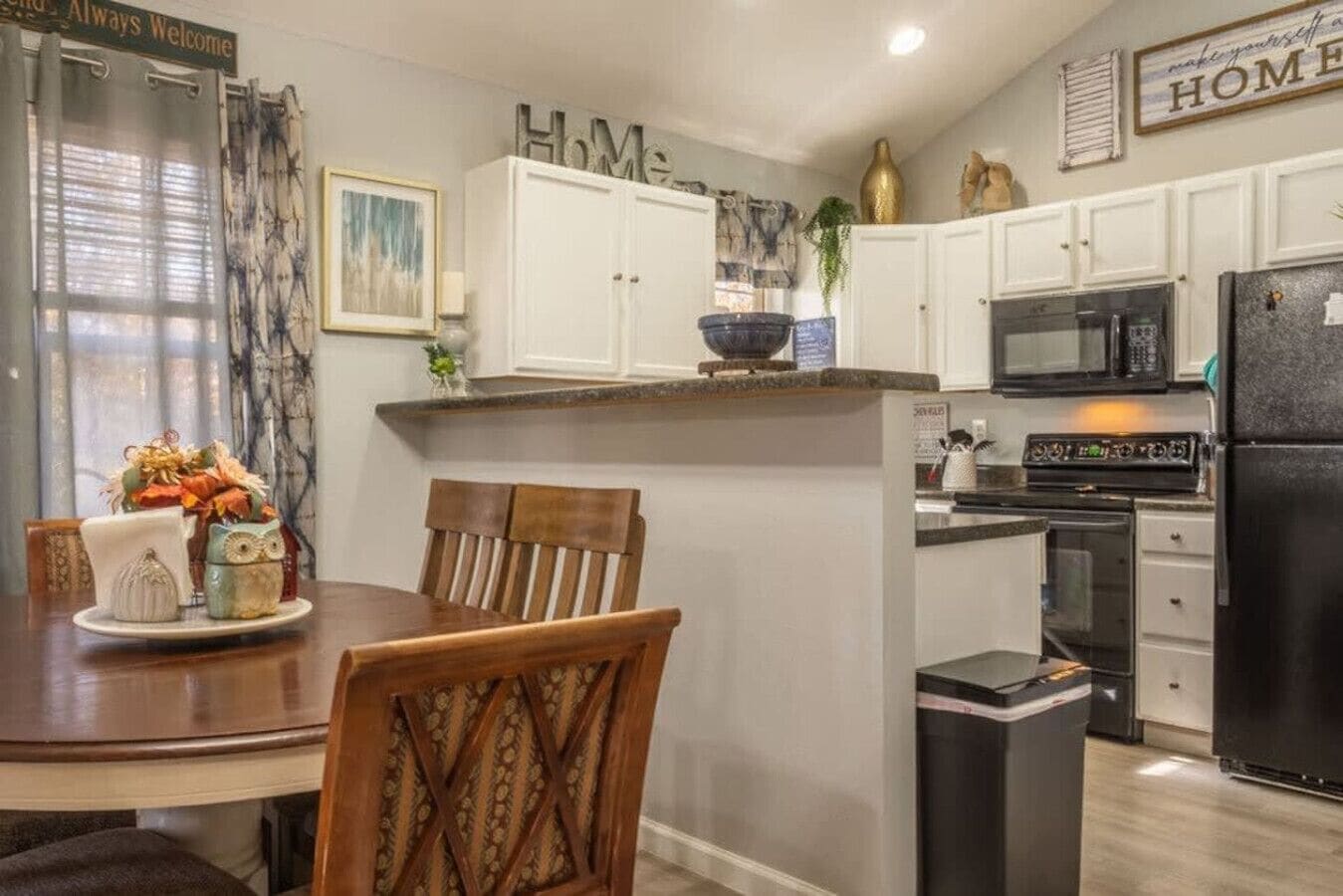 Cocina compacta con armarios blancos, un refrigerador negro y un comedor con mesa y sillas de madera.