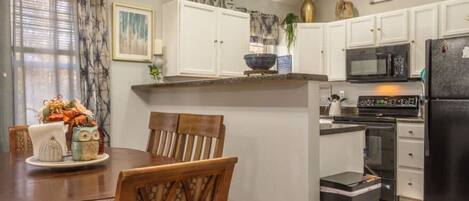 Cocina compacta con armarios blancos, un refrigerador negro y un comedor con mesa y sillas de madera.