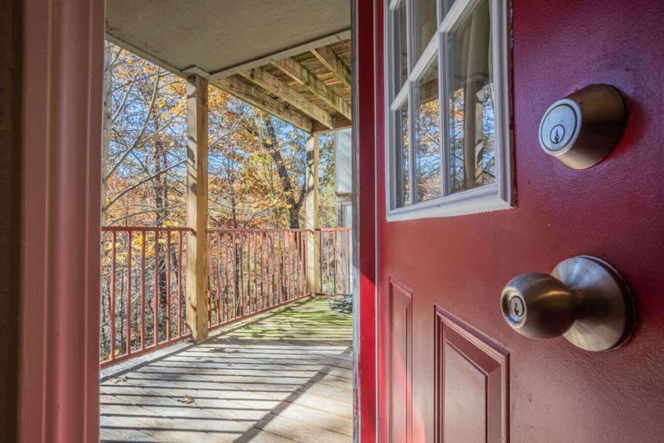 A wooden deck with a metal railing, a red door with a keyhole, and a view of autumn trees.