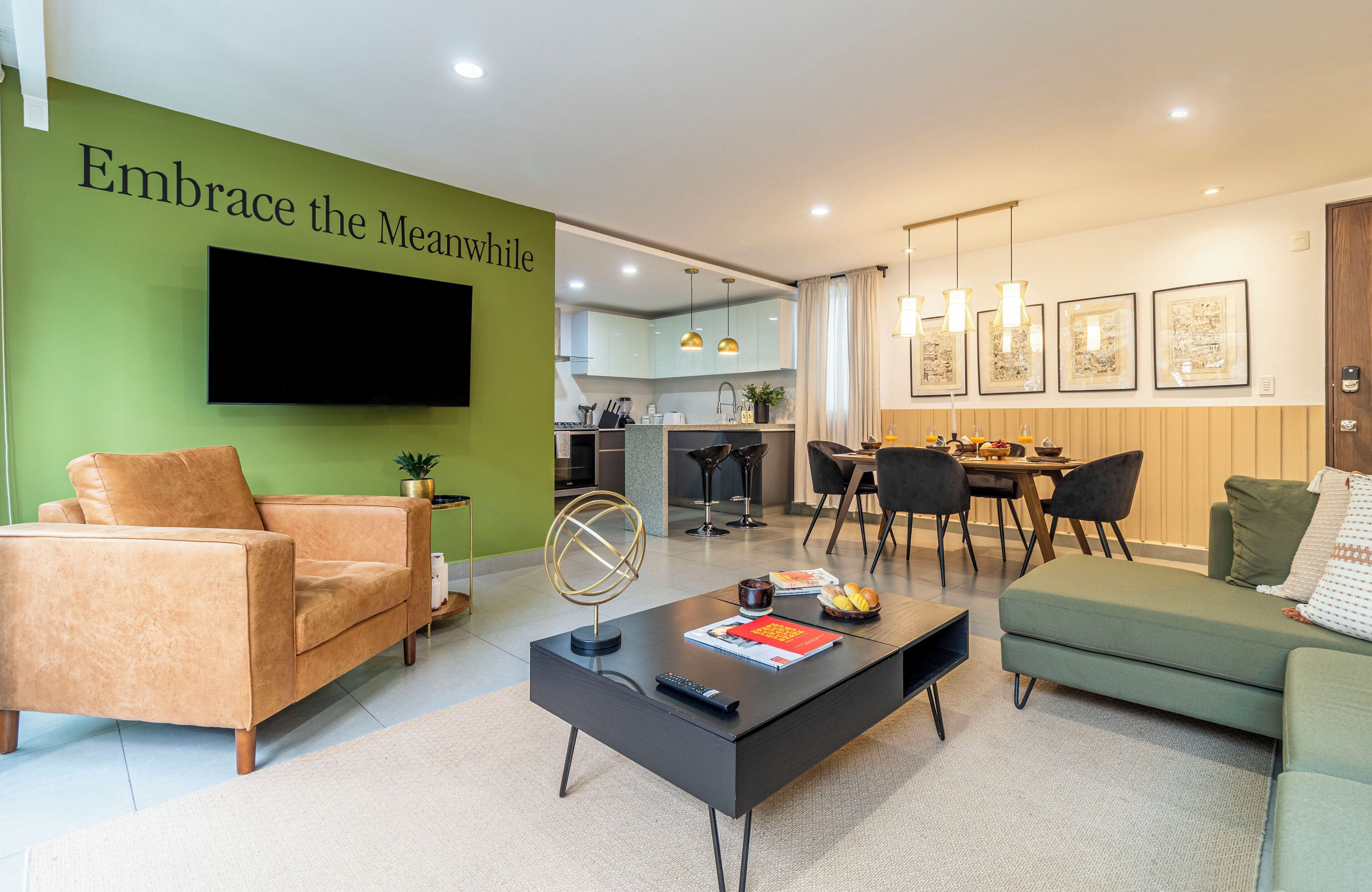 A modern living room with a green accent wall, a flat-screen TV, a coffee table with remote and book, and a dining area with chairs and a table.