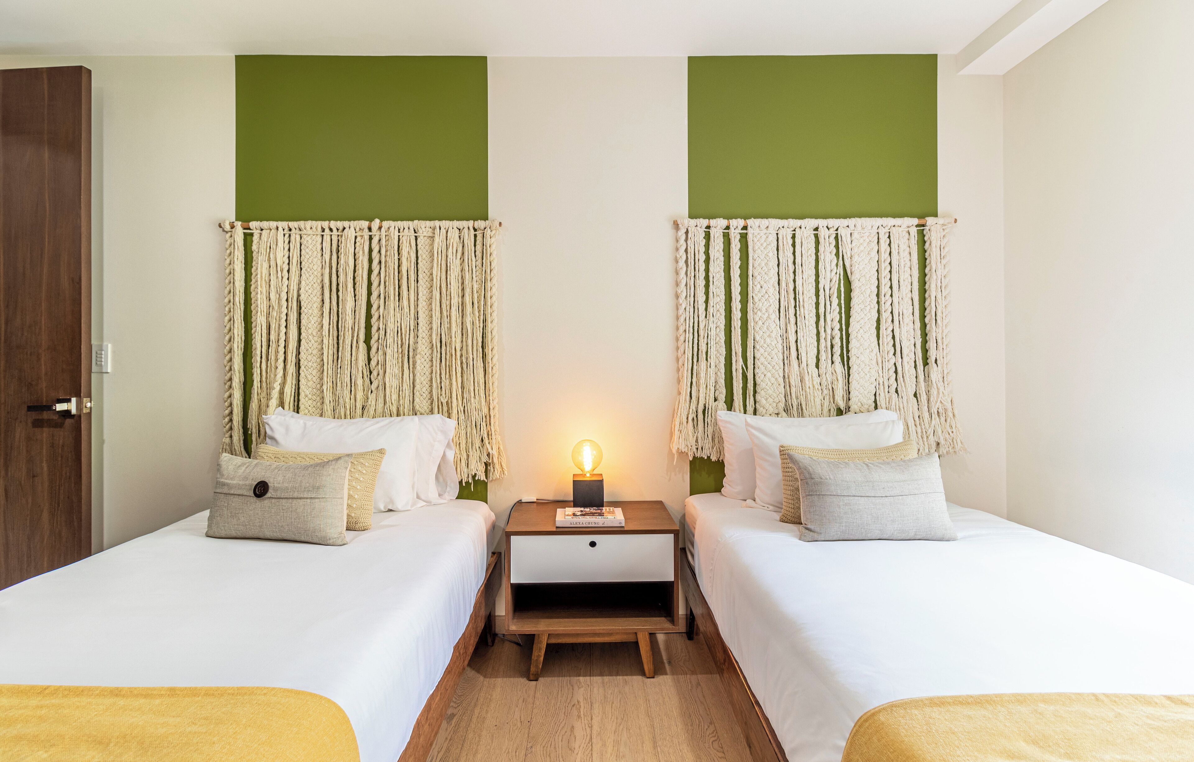 A room with two beds, a nightstand with a lamp, and macramé wall hangings.