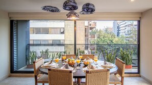 Un comedor con una mesa redonda, sillas de mimbre y vistas a edificios y árboles a través de amplios ventanales.