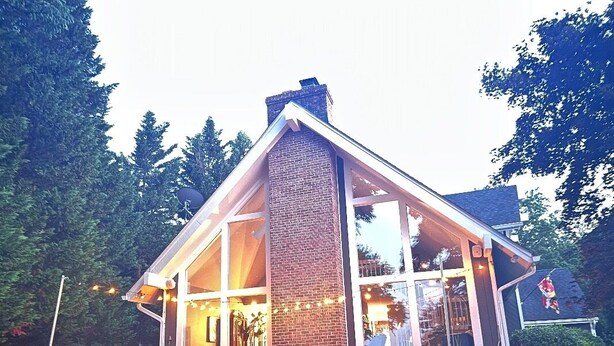 Uma casa acolhedora e moderna, com chaminé de tijolos, janelas grandes e um alpendre com mobiliário para exterior.