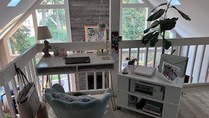 Um escritório em casa com uma mesa, cadeira e estante. Há janelas que oferecem luz natural e vista para o exterior.