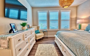 A bedroom with a bed, dresser, chair, windows, and a television.