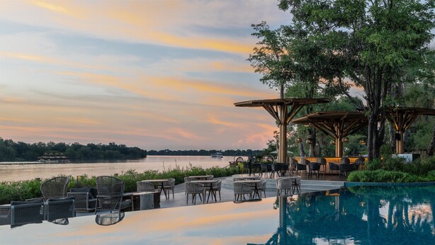 A serene outdoor pool area with lounge chairs and tables, overlooking a calm body of water and a sunset sky.