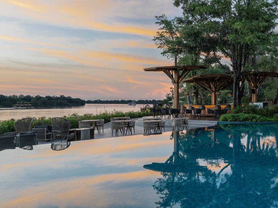 Área de piscina exterior tranquila con sillones y mesas, con vistas a un cuerpo de agua en calma y un cielo al atardecer.