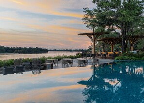 Un espace extérieur paisible avec une piscine, des chaises longues et des tables, offrant une vue sur un plan d’eau calme et un ciel au coucher du soleil.