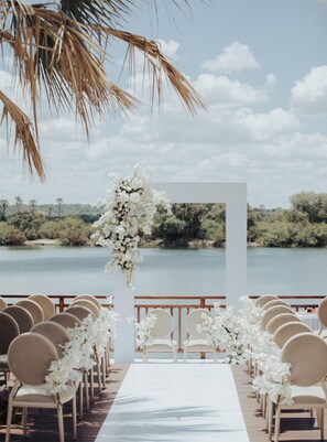 A wedding ceremony setup with a white aisle, chairs, and floral decorations against a lake backdrop.