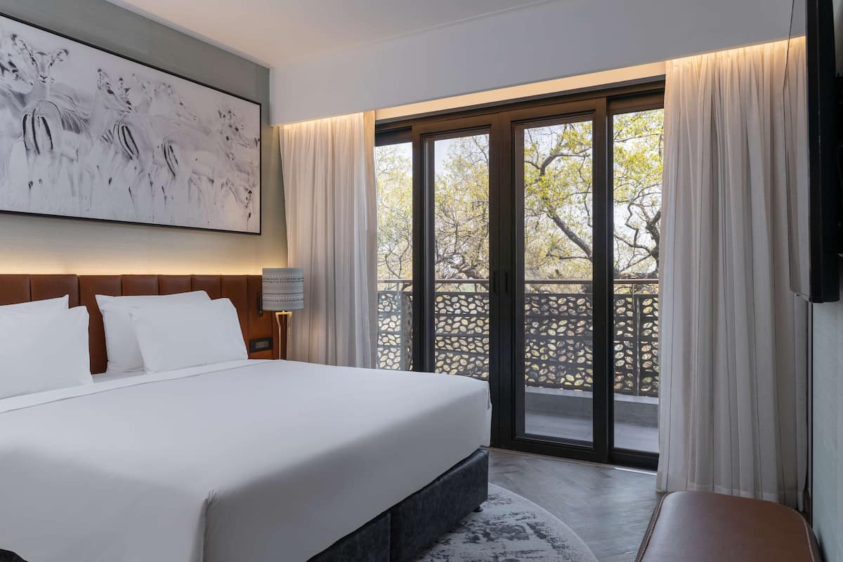 Une chambre d’hôtel avec un grand lit, une tête de lit dotée d’un luminaire, un tableau au mur et un balcon offrant une vue sur les arbres.