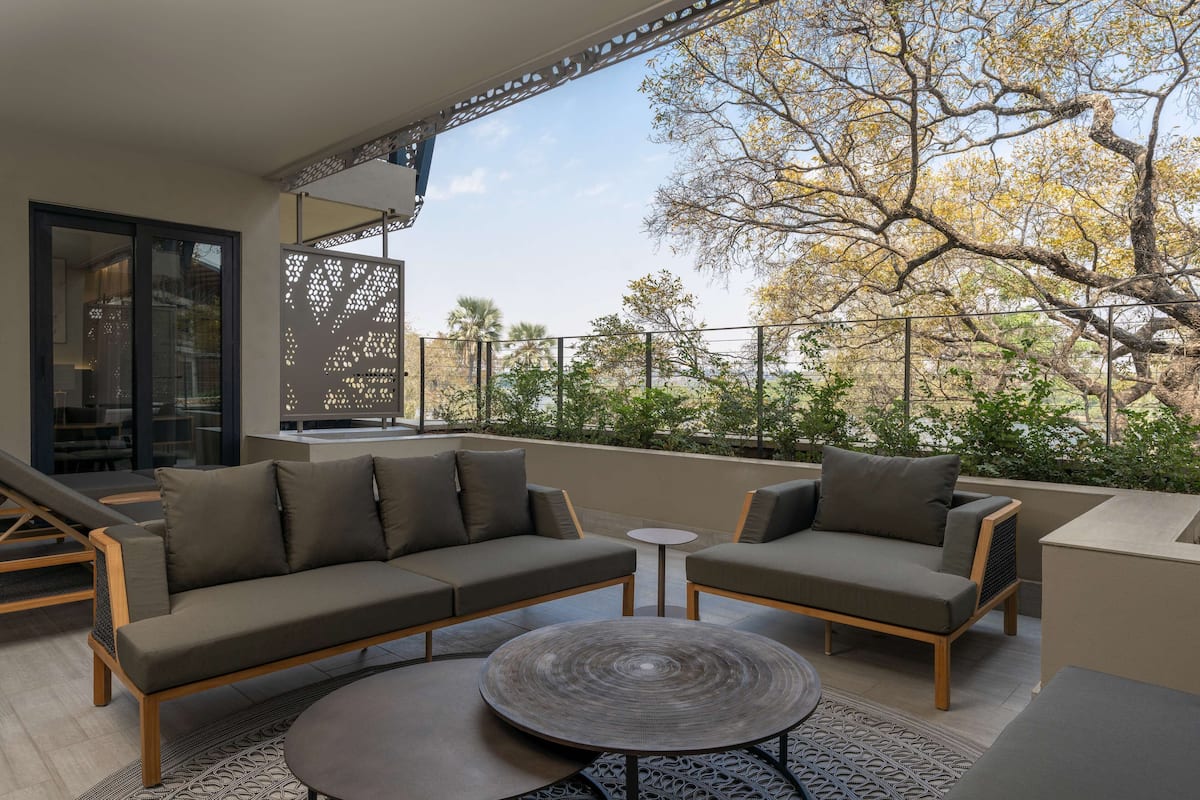 Un espace extérieur moderne aménagé avec un canapé, deux fauteuils et une petite table, donnant sur un jardin avec des arbres et une clôture.