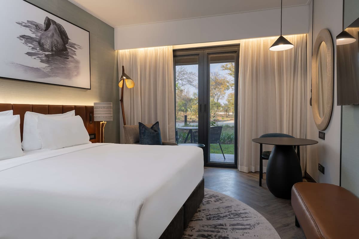 Une chambre d’hôtel moderne, dotée d’un grand lit, d’un bureau, d’une chaise et offrant une vue sur l’extérieur grâce à une porte vitrée.