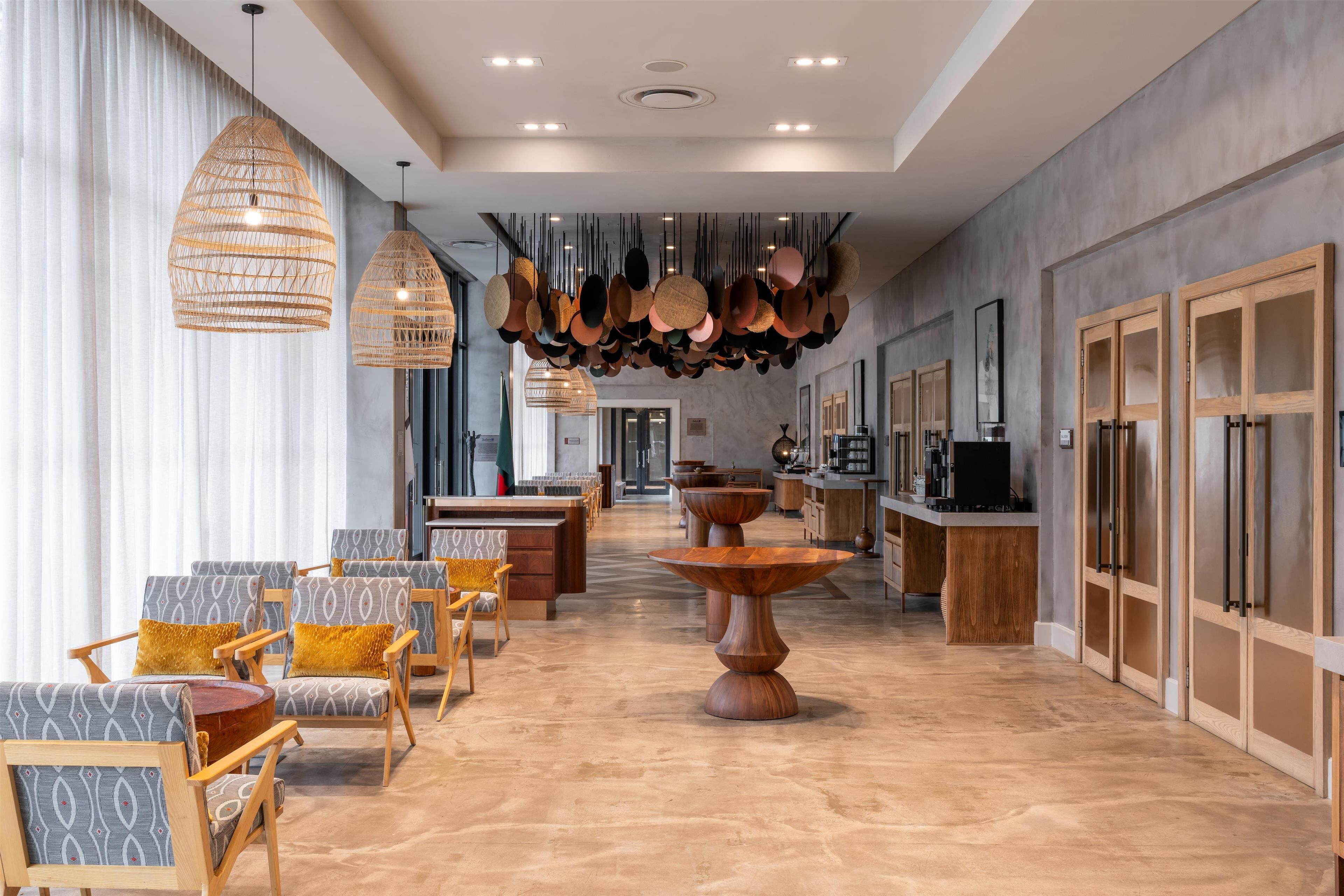 A spacious hotel lobby with modern furniture, a central wooden table, and hanging pendant lights.