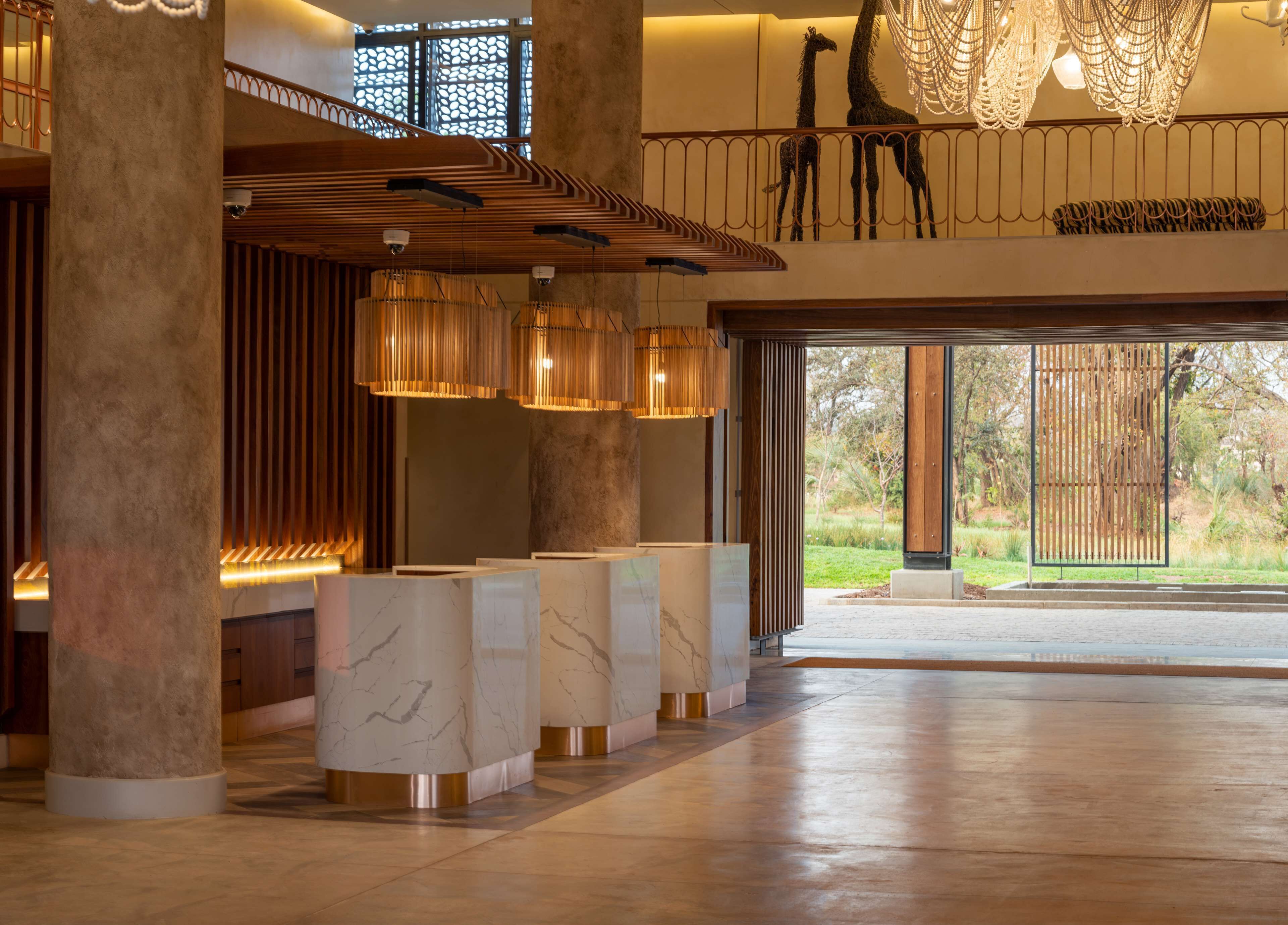 A spacious lobby with marble reception counters, a high ceiling with a wooden slat design, and a view of a garden through large glass doors.