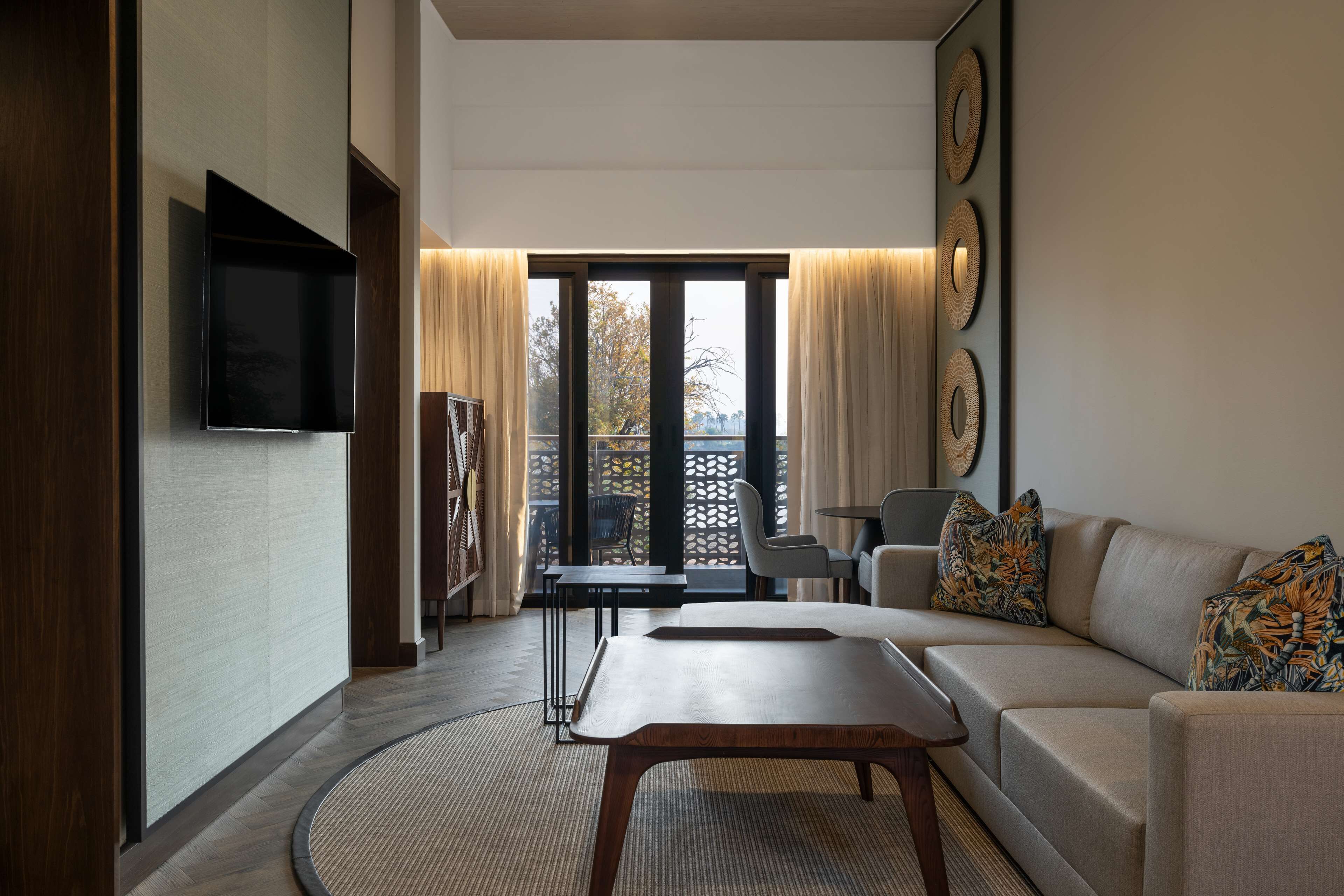 Ein modernes Wohnzimmer mit einer Couch, einem Couchtisch und einem an der Wand befestigten Flachbildfernseher. Ein Balkon bietet Blick auf Bäume und ist mit einem Geländer versehen.
