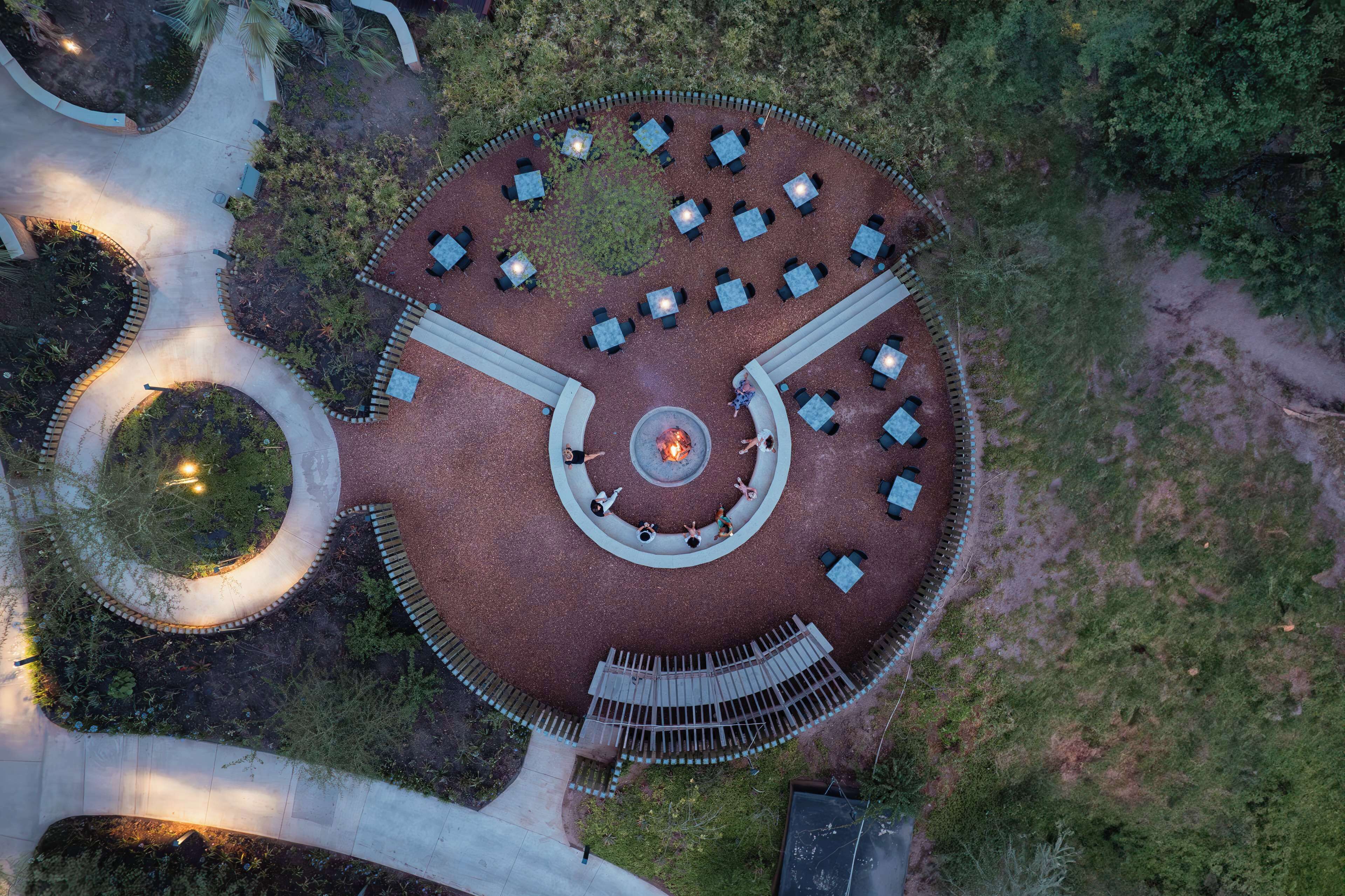 Vista aérea de un espacio de estar circular al aire libre con mesas y sillas, rodeado de un jardín ajardinado.