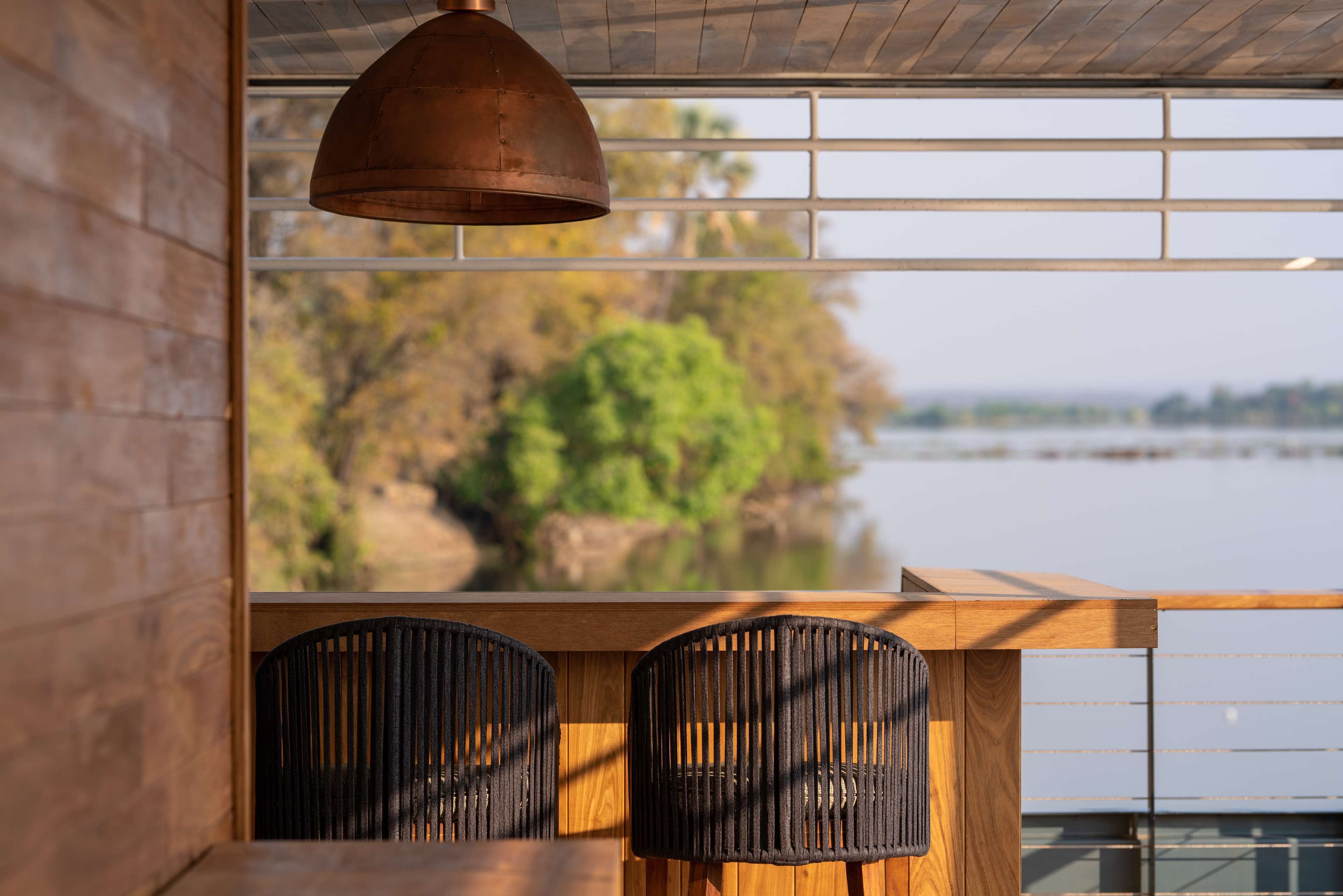 A wooden table with two black chairs, a hanging copper pendant light, and a view of a river and trees through a window with horizontal blinds.