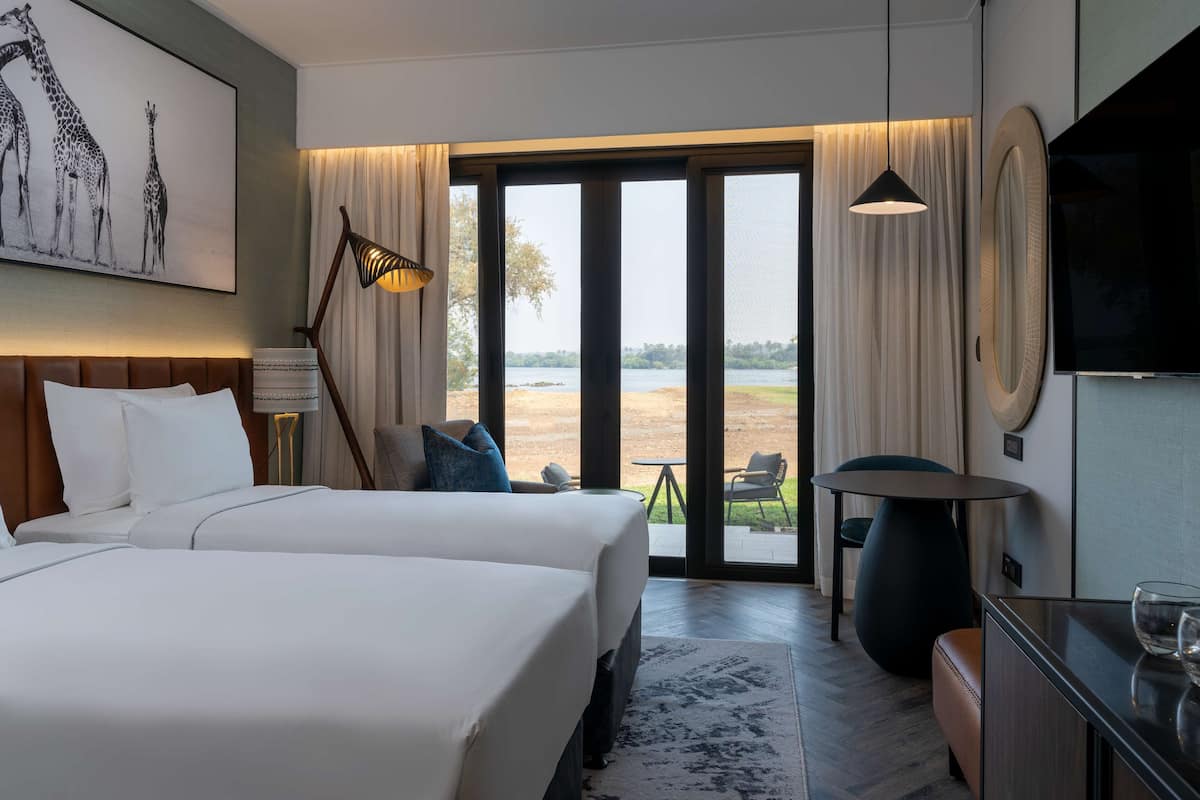 Une chambre d’hôtel avec deux lits, une grande fenêtre donnant sur l’extérieur, une télévision et un coin repas avec une table et des chaises.