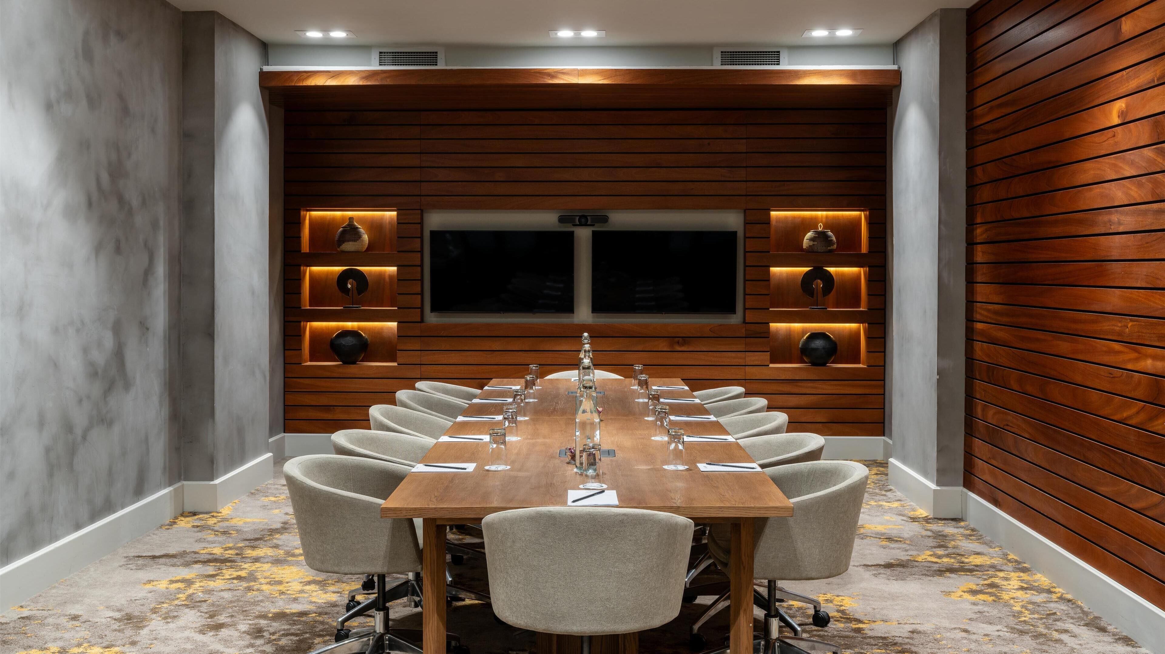 A conference room with a long table, chairs, and a wooden panel wall with built-in lighting.