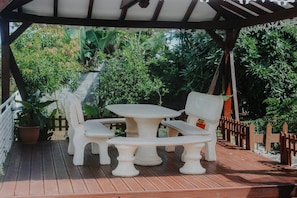 Un patio cubierto con terraza de madera, mobiliario exterior blanco y abundante vegetación al fondo.
