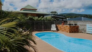 A swimming pool with a fenced area, a building with a green roof, and a sign in the background. - Le Lodge Phoenix, F2 in residence with swimming pool (Sainte-Rose)